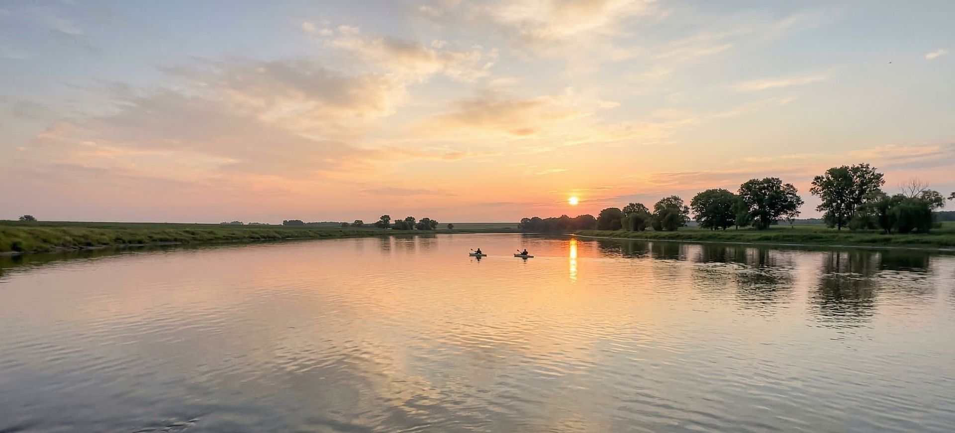Kayakers on a wide heartland river at sunset with flat green banks